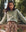 A woman leans against a stone ledge, surrounded by trees, dressed in THE GREAT. SP25 D1's The League Sweatshirt in smoke green with a bucking bronco graphic and paired with a floral skirt.