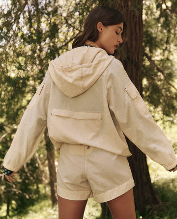 A woman stands outdoors wearing The Trailhead Pullover by THE GREAT. SP25 TGO, a cream hooded jacket paired with matching shorts amidst lush greenery—ideal outdoor wear for hiking enthusiasts.