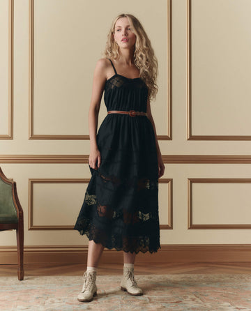 A woman wears THE GREAT. SP26 D2 The Rosette Crochet Dress in black, styled with a brown belt and white ankle boots, standing in a classic room with beige paneled walls and a green chair.