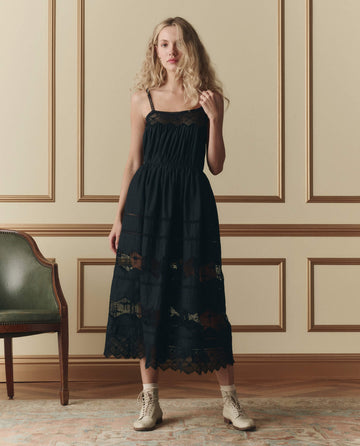 A woman stands indoors wearing THE GREAT. SP26 D2 The Rosette Crochet Dress in black, styled with light-colored lace-up boots; a green chair is beside her.