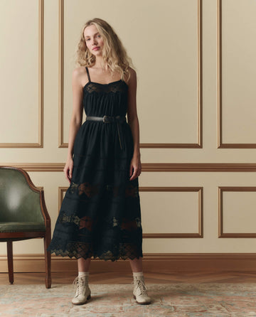 A woman stands indoors by a green chair, wearing THE GREAT. SP26 D2 The Rosette Crochet Dress in black, styled with a belt and light ankle boots against a paneled wall.