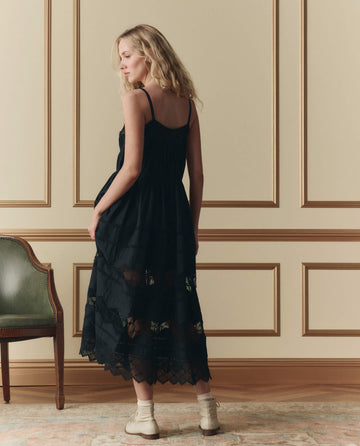 A woman stands sideways by a paneled wall and green chair, wearing THE GREAT. SP26 D2 The Rosette Crochet Dress in black, styled with white socks and beige shoes.