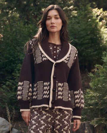 A woman stands outdoors in The Chalet Cardigan Sweater. -- Walnut And Cream by THE GREAT. F26 TGO, surrounded by green foliage and trees.