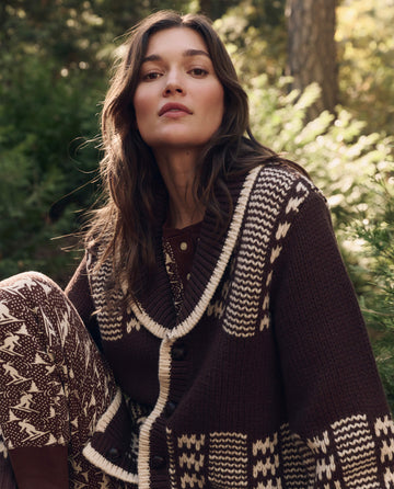 A woman with long brown hair wears THE GREAT. F26 TGO The Chalet Cardigan Sweater. -- Walnut And Cream, sitting outdoors among lush green foliage.