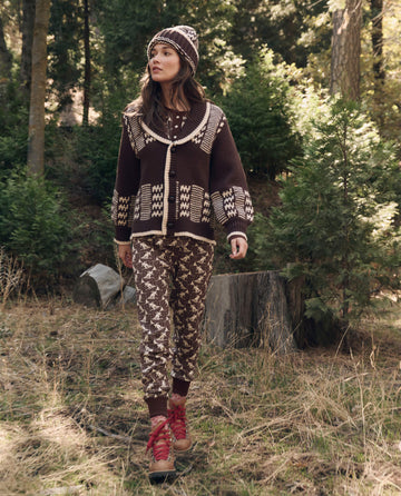 A woman in The Chalet Cardigan Sweater. -- Walnut And Cream by THE GREAT. F26 TGO and red boots walks through a forest clearing among trees and greenery.