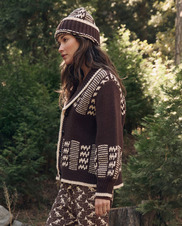 A woman stands outdoors in lush greenery wearing THE GREAT. F26 TGO The Chalet Cardigan Sweater. -- Walnut And Cream, with a matching knit hat and pants.