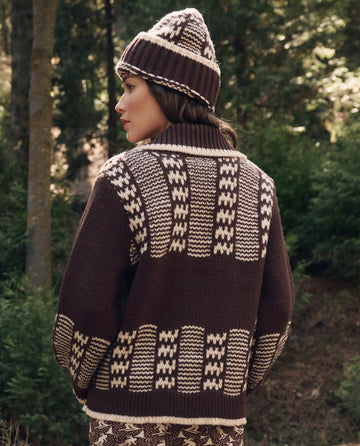 A person outdoors among trees wears The Chalet Cardigan Sweater. -- Walnut And Cream by THE GREAT. F26 TGO and a matching hat.