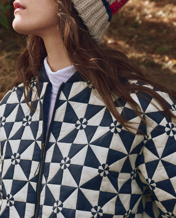 A person wearing THE GREAT. F25 TGO The Alps Puffer Jacket in Quilted Vintage Navy with a zipper and a knit beanie, photographed outdoors.