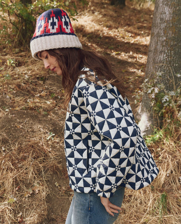 A person wearing THE GREAT. F25 TGO The Alps Puffer Jacket in Quilted Vintage Navy, blue jeans, and a knitted hat stands outdoors near trees and dry grass.
