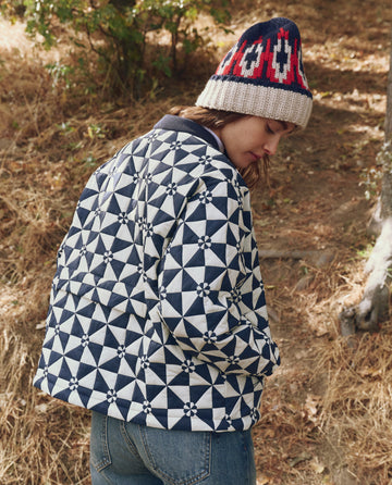 Wearing THE GREAT. F25 TGO's The Alps Puffer Jacket in Quilted Vintage Navy, a person stands outdoors on dry grass, facing away and looking down, paired with jeans and a knit hat.