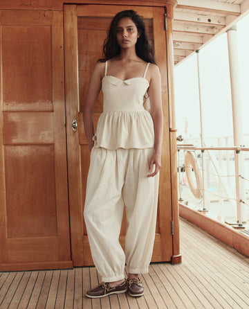A woman stands on a wooden deck wearing THE GREAT. SU26 The Verse Top in Natural with thin straps, matching loose pants, and brown boat shoes.
