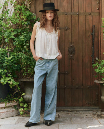 A person stands in front of a wooden door surrounded by green plants, wearing THE GREAT. PF25 The Mill Jean in Dakota Wash, paired with a black hat and a white sleeveless top.