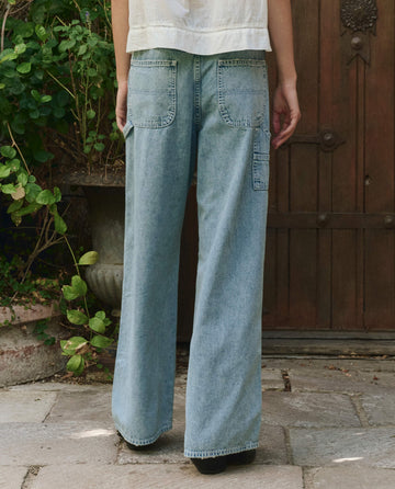 Person stands outdoors on a stone path, wearing THE GREAT. PF25 The Mill Jean in Dakota Wash (light blue wide-leg denim) and a white top, facing away from the camera with arms relaxed by their sides.