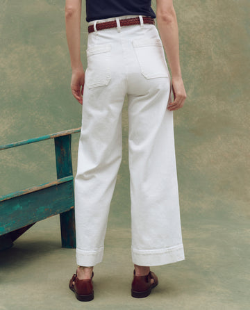 A person stands facing away, wearing THE GREAT. SP25 D1 The Seafair Jean in True White, styled with a brown belt and brown shoes, one hand resting by their side.