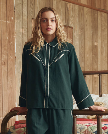 A woman with wavy blonde hair stands indoors, wearing THE GREAT. H25 SLEEP's The Flannel Western Pajama Shirt in Pine & Cream, leaning on a metal bed frame against a wooden wall.