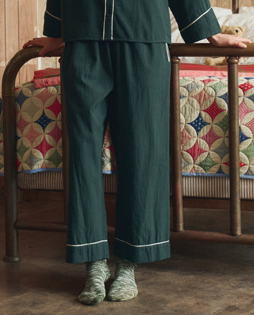 Someone wearing THE GREAT. H25 SLEEP's Flannel Western Easy Sleep Pant in Pine & Cream stands before a metal bed topped with a colorful quilt, their patterned socks adding extra style.