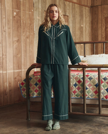 A person in THE GREAT. H25 SLEEP Flannel Western Easy Sleep Pant in Pine & Cream stands at the foot of a metal bed with a colorful patchwork quilt in a wood-paneled room.