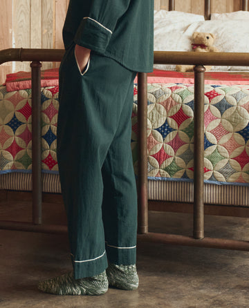 A person wearing THE GREAT. H25 SLEEP's Flannel Western Easy Sleep Pant in Pine & Cream stands beside a vintage metal bed topped with a colorful patchwork quilt and a teddy bear.