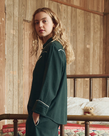 A person with long, wavy hair stands by a metal-framed bed in a wood-paneled room, wearing THE GREAT. H25 SLEEP’s The Flannel Western Pajama Shirt in Pine & Cream. A teddy bear rests on the bed in the background.
