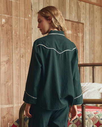 A person with shoulder-length hair, seen from behind in a wood-paneled room by a metal-frame bed, wears THE GREAT. H25 SLEEP’s The Flannel Western Pajama Shirt in Pine & Cream with white piping.