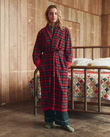 A person is indoors wearing THE GREAT. H25 SLEEP’s The Plush Fleece Robe in Cedarwoods Plaid over dark pajamas, with a wooden bed and patchwork quilt behind them.