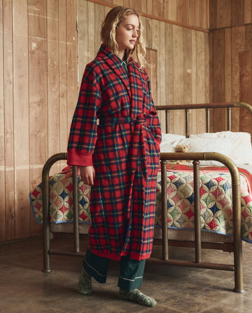 A woman stands by a brass bed in a wood-paneled room, wrapped in THE GREAT. H25 SLEEP The Plush Fleece Robe – Cedarwoods Plaid, wearing dark green pajamas and green socks.