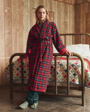 Wearing THE GREAT. H25 SLEEP’s The Plush Fleece Robe in Cedarwoods Plaid, a person stands by a brass bed with a patchwork quilt in a warmly wood-paneled room.