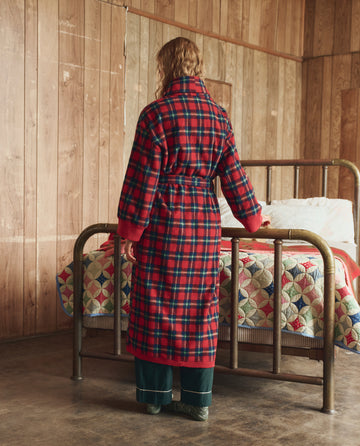 A person wears The Plush Fleece Robe in Cedarwoods Plaid by THE GREAT. H25 SLEEP, standing at the foot of a brass bed in a wood-paneled room.
