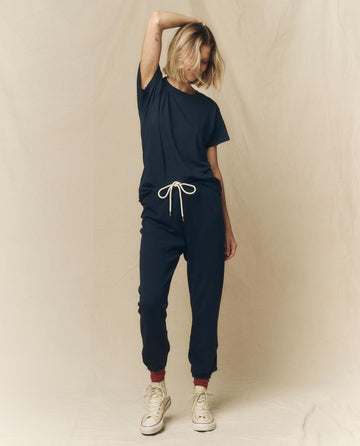 A person stands with one arm overhead, wearing THE GREAT. FALL 23 KNITS' The Slim Tee in True Navy, paired with drawstring pants made in the U.S.A., white sneakers, and red ankle socks.