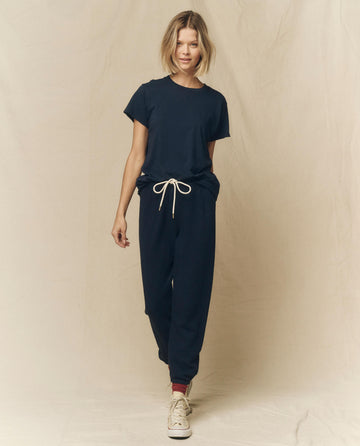 A person with short blonde hair is wearing The Slim Tee. Solid in True Navy by THE GREAT. FALL 23 KNITS, along with sweatpants featuring a white drawstring, paired with white sneakers and red socks, standing against a neutral background.