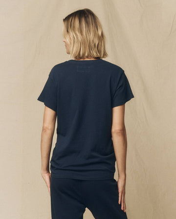 A person with short blond hair facing away is wearing "The Slim Tee" in True Navy by THE GREAT. from their FALL 23 KNITS collection and dark pants, standing against a beige background.