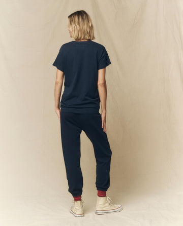 Person standing with their back to the camera, wearing The Slim Tee in True Navy from THE GREAT. FALL 23 KNITS, black sweatpants with red cuffs, and beige sneakers. The background is a plain beige fabric.