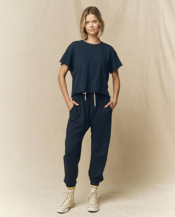 A person stands against a beige backdrop wearing THE GREAT. FALL 23 KNITS' The Crop Tee in True Navy and matching joggers made from super soft cotton, with hands in pockets, and white high-top sneakers.