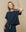 A person with shoulder-length blonde hair wearing THE GREAT. FALL 23 KNITS' "The Crop Tee. Solid -- True Navy" and matching high-rise jeans poses in front of a plain beige background.