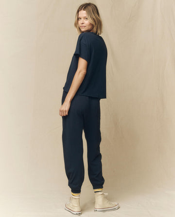 A blonde woman in "The Crop Tee, Solid -- True Navy" from THE GREAT. FALL 23 KNITS and matching jogger pants stands facing away, looking over her shoulder. She is wearing white high-top sneakers with yellow and black striped socks. Her super soft cotton outfit complements her relaxed stance perfectly.