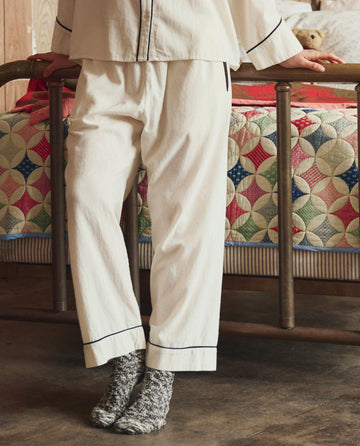 Wearing THE GREAT. H25 SLEEP’s Flannel Western Easy Sleep Pant in Cream & Black, a person stands by a metal bed topped with a colorful patterned quilt.