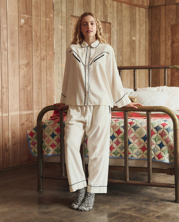 A person in THE GREAT. H25 SLEEP's Flannel Western Easy Sleep Pant in Cream & Black, with socks, stands by a metal bed with a colorful quilt in a wood-paneled room with a concrete floor.