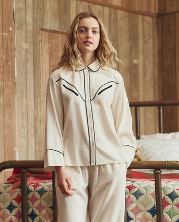 A person with long wavy hair stands indoors by a wooden wall and patchwork quilt, wearing THE GREAT. H25 SLEEP’s The Flannel Western Pajama Shirt in Cream & Black, featuring black piping details.