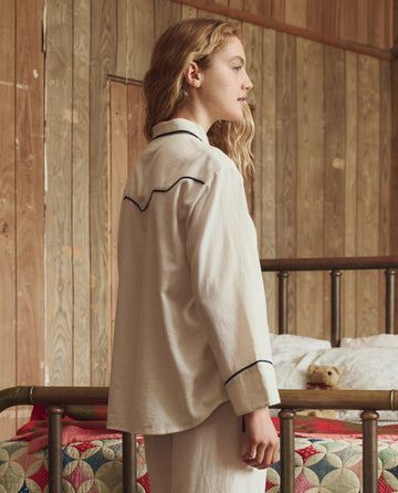 A person with long hair stands in profile beside a metal bed in a wood-paneled room, wearing THE GREAT. H25 SLEEP's The Flannel Western Pajama Shirt in Cream & Black.
