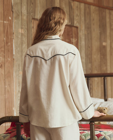 A person with long wavy hair, seen from behind, wears THE GREAT. H25 SLEEP's The Flannel Western Pajama Shirt in Cream & Black near a bed in a wood-paneled room.