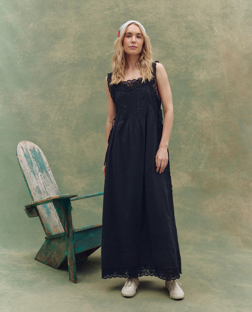 A woman in THE GREAT. SU25 The Attic Lace Dress in black, white sneakers, and a light headscarf stands by a rustic green wooden chair against a textured green backdrop.