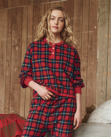 A person wears THE GREAT. H25 SLEEP's Plush Fleece Henley Sleep Sweatshirt in Cedarwoods Plaid, standing indoors by a wood-paneled wall with a bed partially visible beside them.