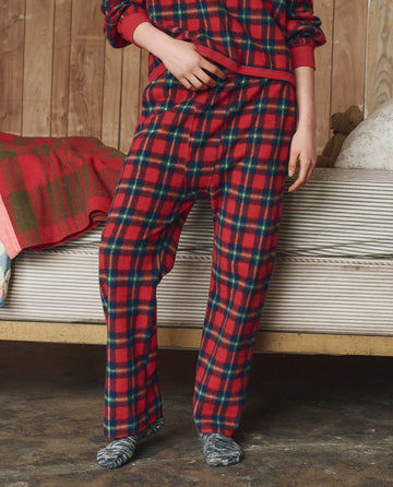A person wearing THE GREAT. H25 SLEEP The Plush Fleece Pajama Pant in Cedarwoods Plaid stands beside a bed with a plaid blanket in a wood-paneled room.