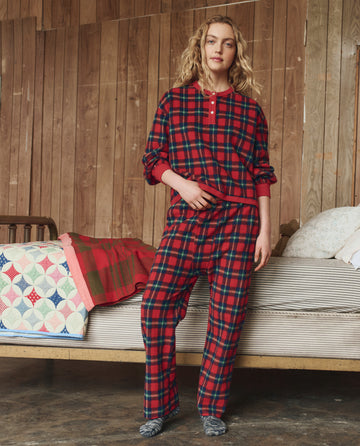 A person stands indoors in THE GREAT. H25 SLEEP The Plush Fleece Pajama Pant – Cedarwoods Plaid, holding the matching shirt. Behind them is a bed with quilts and pillows against a wooden wall.