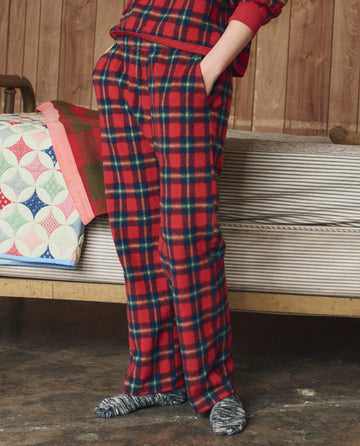 A person wearing THE GREAT. H25 SLEEP’s The Plush Fleece Pajama Pant in Cedarwoods Plaid stands indoors beside a bed with a colorful quilt.