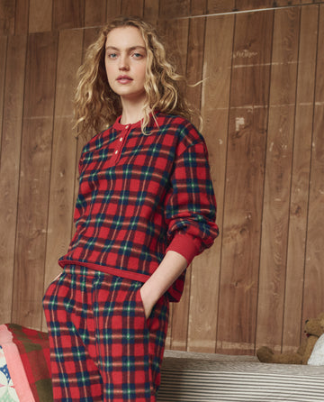 A person with long curly hair stands indoors before a wooden wall, wearing THE GREAT. H25 SLEEP's Plush Fleece Henley Sleep Sweatshirt in Cedarwoods Plaid.