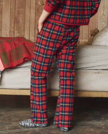 A person stands indoors in THE GREAT. H25 SLEEP The Plush Fleece Pajama Pant in Cedarwoods Plaid, paired with socks, by a bed with a striped blanket and teddy bear.