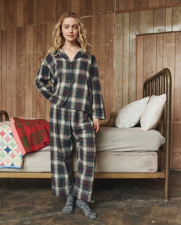 A person stands beside a bed in a wooden room, wearing THE GREAT. H25 SLEEP The Flannel Easy Sleep Pant in Pine Lodge Plaid. The bed is made with striped bedding, pillows, and two folded blankets.
