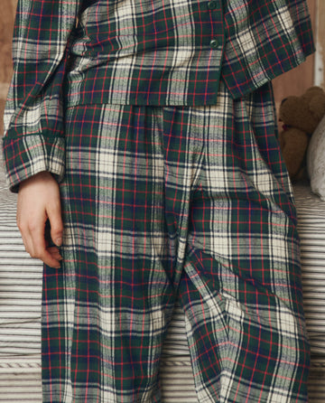 Someone wearing THE GREAT. H25 SLEEP The Flannel Easy Sleep Pant in Pine Lodge Plaid stands next to a bed with a striped blanket, with only their torso and legs visible.