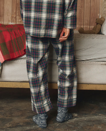 A person stands indoors next to a bed with a striped blanket and teddy bear, wearing THE GREAT. H25 SLEEP The Flannel Easy Sleep Pant in Pine Lodge Plaid and patterned socks, facing away from the camera.